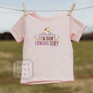 May include: Pink short-sleeved t-shirt with a brown and white cowboy hat, horseshoes, and flowers. The text on the shirt reads "can't talk right now I'M DOIN' COWGIRL STUFF paws-itivi-tee".