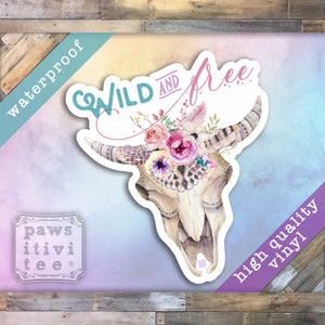 May include: A white cow skull with a floral crown of pink, purple, and white flowers. The skull has a tribal design and the text "WILD AND FREE" is written in pink and teal.