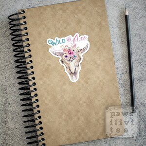 May include: A sticker with a watercolor illustration of a cow skull with a floral crown. The text "Wild and Free" is written above the skull in a turquoise and pink font.