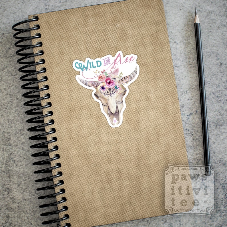May include: A sticker with a watercolor illustration of a cow skull with a floral crown. The text "Wild and Free" is written above the skull in a turquoise and pink font.