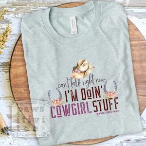 May include: A light blue t-shirt with a graphic design of a cowboy hat, horseshoes, and flowers. The text on the shirt reads "can't talk right now I'M DOIN' COWGIRL STUFF" and "paws-itivi-tees".