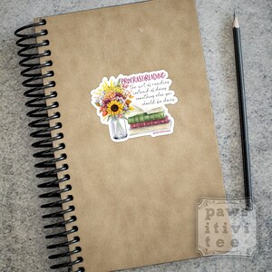 Pu&ograve; includere: Un taccuino a spirale con copertina color marrone chiaro e una matita nera, con un adesivo raffigurante un vaso di vetro pieno di girasoli e fiori autunnali accanto a una pila di libri con il testo "PROCRASTIREADING: the art of reading instead of doing something else you should be doing."