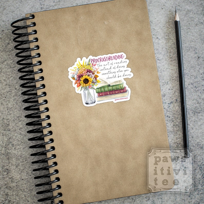 Pu&ograve; includere: Un taccuino a spirale con copertina color marrone chiaro e una matita nera, con un adesivo raffigurante un vaso di vetro pieno di girasoli e fiori autunnali accanto a una pila di libri con il testo "PROCRASTIREADING: the art of reading instead of doing something else you should be doing."