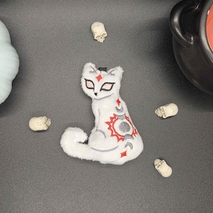 May include: A white plush cat ornament featuring embroidered red sun and grey moon phase designs on its back, with pointed ears and almond-shaped eyes. The cat is surrounded by a light blue pumpkin-shaped candle holder and four small skulls.