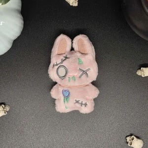 May include: A small, light pink plush rabbit toy featuring embroidered details including a circular eye patch, an X-shaped eye, stitched scars, a small blue flower on the torso, and green accents. The rabbit has long ears and a soft, fuzzy texture.