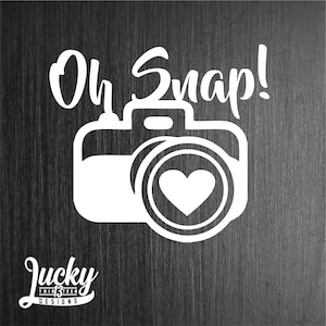 May include: White vinyl decal with a camera silhouette and a heart in the lens. The text "Oh Snap!" is above the camera. The text "Lucky Thirteen Designs" is below the camera.