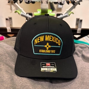 Può includere: Cappellino da camionista nero con una toppa che recita "NEW MEXICO" in lettere gialle, con un bordo turchese e un simbolo. La toppa dice anche "ESTABLISHED 1912". Il cappellino ha un'etichetta Richardson 112 Trucker.