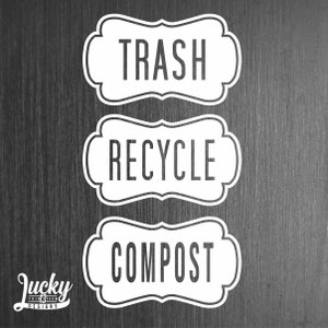 May include: Three white labels with black text on a dark wood background. The labels are shaped like a banner with rounded corners. The labels read "Trash", "Recycle", and "Compost".