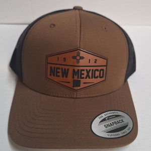 May include: A brown and black trucker hat with a brown leather patch that says "1912 New Mexico" with a symbol of the state of New Mexico. The hat has a black mesh back and a brown brim. The hat has a silver sticker on the bottom of the brim that says "The Authentic Yupoong Snapback Yupoong Since 1974."