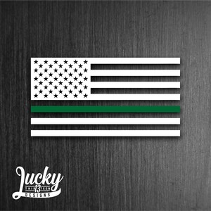 May include: A white American flag with a thin green stripe across the middle. The flag is on a dark wood background. The text "Lucky Thirteen Designs" is below the flag.