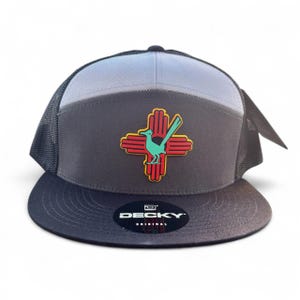 May include: A trucker hat with a gray and light blue panel design. The hat features a patch with a roadrunner bird and a red and yellow Zia symbol. The brim and mesh backing are black. The brand name "DECKY" is printed on the front.