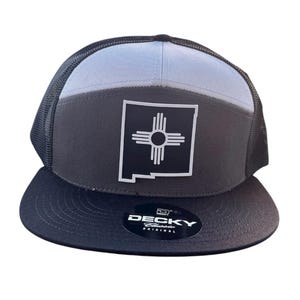Può includere: Cappellino da camionista nero e grigio con il simbolo dello stato del New Mexico. Il cappellino ha una visiera nera e un retro in rete, con un pannello frontale grigio e nero. Il simbolo è un sole Zia bianco all'interno di un quadrato nero.