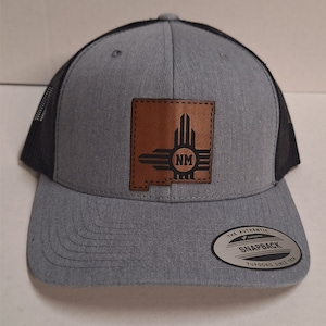 Op de afbeelding: Een grijze en zwarte truckercap met een bruin leren patch met de staat New Mexico en de letters "NM". De pet heeft een zwart mesh achterkant en een grijze voorkant met een gebogen klep. De pet is gelabeld "The Authentic Yupoong Snapback Yupoong Since 1974".