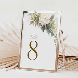 Könnte beinhalten: Eine silbergerahmte Tischnummerkarte mit floralem Design. Die Karte zeigt eine Aquarellillustration von weißen und grünen Hortensien und Blättern. Die Nummer "8" wird in Gold angezeigt, mit dem Wort "Table" darüber.