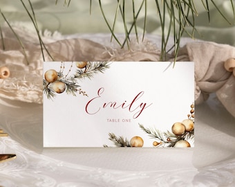May include: A white place card with a watercolor illustration of pine branches and gold berries. The name "Emily" is written in red cursive script, and the text "Table One" is printed in red below the name.
