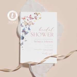 Op de afbeelding: Een witte bruidsdouche-uitnodiging met een roze en blauwe aquarel-vlinderontwerp. De uitnodiging luidt "bridal shower" in roze schrijfletter. De tekst bevat de naam van de bruid, de datum, tijd en locatie van de douche. De uitnodiging bevat ook de zin "kindly reply" in roze schrijfletter.
