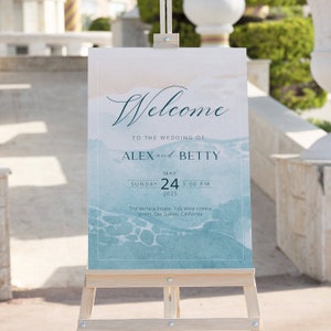 以下が含まれることがあります： 青い水彩画の背景に「AlexとBettyの結婚式へようこそ」という文字が書かれた、結婚式用のウェルカムサイン。サインは木製イーゼルに置かれています。