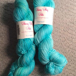 May include: Two skeins of teal blue yarn with white labels that read "Stunning String Studio" and "Tranquil".