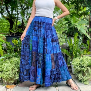 Op de afbeelding: Lange patchwork rok in blauwtinten, met diverse patronen en ontwerpen. De rok wordt gecombineerd met een witte mouwloze top. Het ontwerp van de rok omvat olifantenmotieven en andere ingewikkelde details, wat een bohemien esthetiek creëert.