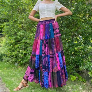 Puede incluir: Una falda larga de patchwork con una vibrante mezcla de colores que incluye rosa, azul y morado. La falda tiene un estilo bohemio con varios patrones y dise&ntilde;os, incluyendo motivos de elefantes. La falda se combina con una camiseta blanca de manga corta.