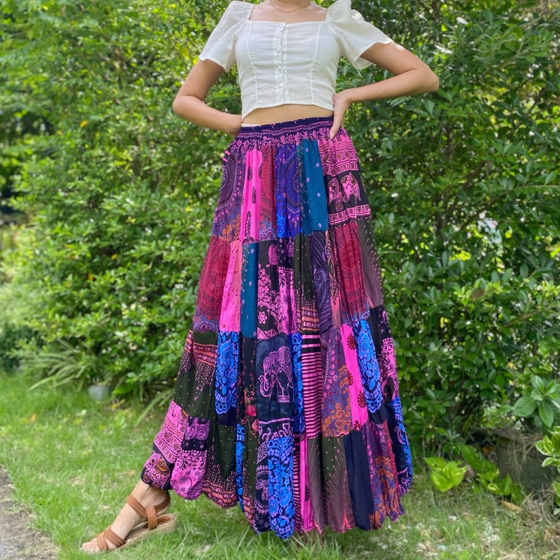 Puede incluir: Una falda larga de patchwork con una vibrante mezcla de colores que incluye rosa, azul y morado. La falda tiene un estilo bohemio con varios patrones y dise&ntilde;os, incluyendo motivos de elefantes. La falda se combina con una camiseta blanca de manga corta.