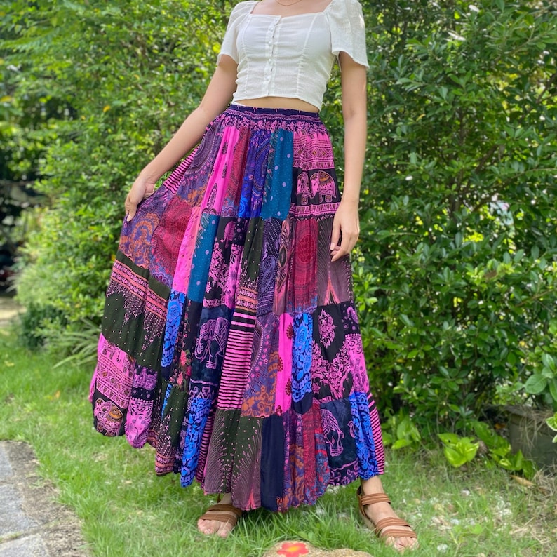 Puede incluir: Falda larga de patchwork con una mezcla vibrante de colores como rosa, morado, azul y negro. La falda tiene un dise&ntilde;o escalonado con varios paneles de tela estampada. Se combina con una blusa blanca y sandalias marrones, creando un estilo bohemio.