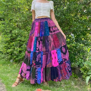 Puede incluir: Una falda larga de patchwork en tonos rosas, azules y morados. La falda presenta varios cuadrados de tela estampada, creando un estilo bohemio. La falda se lleva con una camiseta blanca de manga corta.