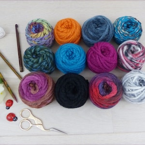 Könnte beinhalten: Eine Sammlung bunter Wollknäuel in verschiedenen Farbtönen, darunter Blau, Orange und Lila, angeordnet auf einer hellen Holzoberfläche. Stricknadeln, ein Häkelhaken, eine Schere und dekorative Marienkäfer deuten auf ein Bastelthema hin.