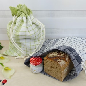 Könnte beinhalten: Zwei Stoffbeutel zum Aufbewahren von Brot. Ein Beutel ist grün-weiß kariert, der andere schwarz-weiß kariert. Beide Beutel haben weiße Kordeln. Ein Laib Brot befindet sich im schwarz-weißen Beutel. Ein kleines Glas mit einem roten und weißen Tupfendeckel befindet sich im Vordergrund.