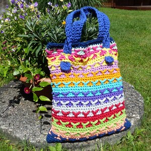 May include: A colourful crocheted tote bag with blue handles and a striped pattern. The bag is made with different coloured yarns, including red, orange, yellow, green, blue, and purple.
