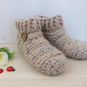 May include: A pair of beige crocheted slippers with a button detail. The slippers have a speckled pattern with hints of purple, pink, and blue.