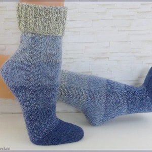 May include: A pair of blue and grey gradient knit socks with a grey ribbed cuff. The socks are shown on a model's feet against a white brick wall background.