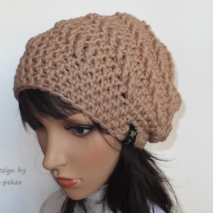 May include: A beige crocheted beanie with a textured design. The beanie has a small black tag with the text "design by p-pekee" on it.