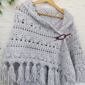 May include: A grey crocheted shawl with a fringe border and a wooden toggle closure. The shawl is draped over a hanger.