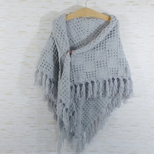 May include: A gray crocheted shawl with fringe hanging from the bottom edge. The shawl is draped over a wooden hanger.