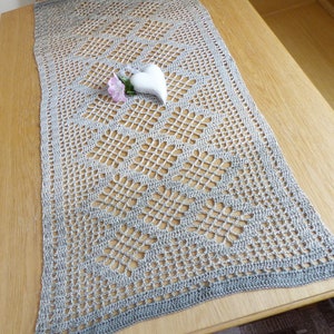 Puede incluir: Un camino de mesa de ganchillo gris con un patrón delicado. El camino de mesa está colocado sobre una mesa de madera con una decoración en forma de corazón blanca en la parte superior.