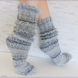 May include: A pair of gray and white knit socks with a ribbed cuff. The socks are folded down to show the ribbed detail. The socks are on a white tiled background.