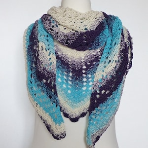 May include: A handmade crochet shawl in shades of cream, teal, and deep purple. The shawl is draped over a mannequin, showcasing its intricate openwork design and scalloped edges. The design is by p-pekee.