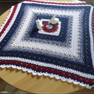 Pode incluir: Um cobertor quadrado de crochê feito à mão em tons de azul marinho, branco e vermelho. O cobertor apresenta uma borda ondulada e um design quadrado central. Duas pequenas figuras de cerâmica branca são colocadas no cobertor.