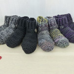 May include: A collection of four pairs of ankle-high crochet slippers arranged in a row. The slippers feature various colors including charcoal gray, solid black, a variegated earth-tone blend, and a deep purple, each accented with a single side button.