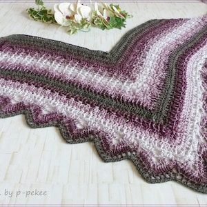 May include: A crocheted shawl in shades of purple, white, and gray with a scalloped edge. The shawl is laid out on a white surface.