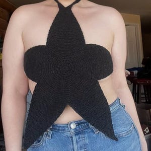 May include: A black, star-shaped crochet top with a halter neck design. The top is made of a textured yarn and is worn with blue denim jeans. The star shape is the focal point of the top.