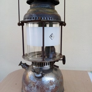 Könnte beinhalten: Eine antike, verwitterte Metalllaterne mit einem durchsichtigen Glaszylinder und einem dunklen, gewölbten Deckel. Die Laterne hat einen Griff und ein kleines Rad an der Seite. Der Glaszylinder hat ein weißes Band mit einem Logo.