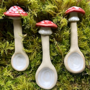 Könnte beinhalten: Drei Keramik-Pilzlöffel mit rot-weiß gepunkteten Kappen und weißen Schalen. Die Löffel sind vor einem Hintergrund aus grünem Moos platziert und schaffen eine skurrile, von der Natur inspirierte Ästhetik. Ideal zum Servieren von Gewürzen.