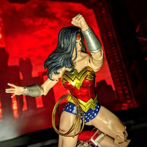 Puede incluir: Figura de acción de Wonder Woman en una pose dinámica, vestida con su icónico traje rojo, dorado y azul. La figura tiene cabello negro y brazaletes plateados, con un lazo dorado enrollado a su lado. El fondo es rojo intenso con una silueta de ciudad.