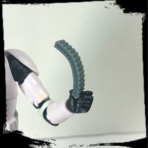 May include: A gray plastic accessory with a curved shape and a series of small, round holes. The accessory is held in a black gloved hand.