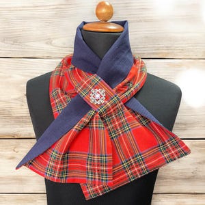 May include: A red plaid scarf with a navy blue collar and accents, displayed on a mannequin. The scarf features a decorative brooch with a pink center. The plaid pattern includes green, yellow, and white lines.