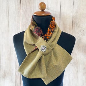 May include: A tan scarf with a decorative brooch. The scarf has a patterned lining with orange, brown, and green accents. The brooch is silver-toned with a central purple stone and surrounding clear stones. The scarf is displayed on a black mannequin.