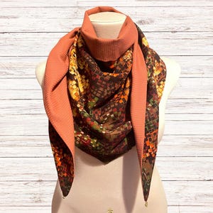 May include: A brown, green, orange, and yellow patterned scarf with a coral pink trim. The scarf is folded and displayed on a mannequin.