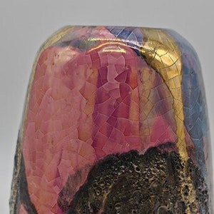 Op de afbeelding: Een kleine, keramische vaas met een uniek, getextureerd oppervlak. De vaas is versierd met een kleurrijke glazuur in tinten roze, goud en blauw.